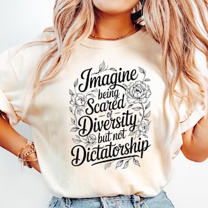 May include: Cream-colored t-shirt with the text "Imagine being Scared of Diversity but not Dictatorship" in a black, floral design. The shirt is paired with light blue denim jeans.