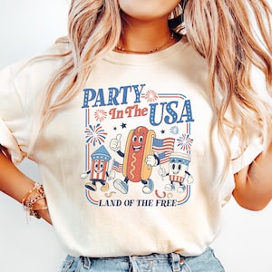 May include: Cream-colored t-shirt with a retro graphic that reads "Party In The USA" in blue and red. The graphic features cartoon characters of a hot dog, a firework, and a cup of soda, all holding American flags. The text "Land of the Free" is below.