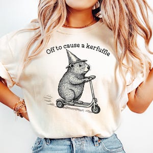 May include: Cream-colored t-shirt with the phrase "Off to cause a kerfuffle." The graphic features a rodent wearing a witch's hat and riding a scooter. The design is in black ink.