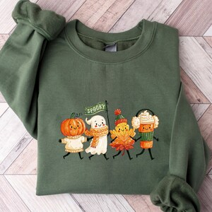 May include: An olive green crewneck sweatshirt featuring a graphic print of four cartoon characters holding hands: a pumpkin, a ghost holding a flag that says SPOOKY, an autumn leaf, and a coffee cup with whipped cream, all wearing winter accessories.