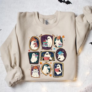 May include: Beige sweatshirt featuring a colorful graphic of cartoon ghosts reading books, each framed in a different ornate style. The ghosts wear hats and scarves, with bats and string lights in the background. A cozy, book-themed design.
