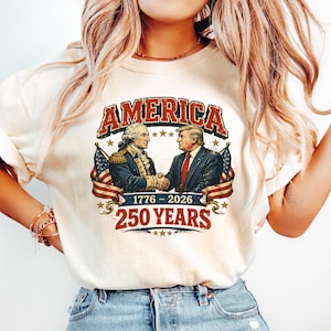 Puede incluir: Camiseta color crema con la palabra "AMERICA" en rojo desgastado. El dise&ntilde;o muestra a dos hombres estrech&aacute;ndose la mano, flanqueados por banderas americanas, con el texto "1776-2026" y "250 YEARS" debajo. La camiseta se combina con vaqueros azul claro.