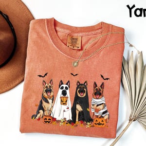 May include: A coral-colored Comfort Colors sweatshirt featuring a Halloween-themed graphic. The design includes four German Shepherds dressed as a ghost, mummy, and other costumes, with pumpkins and bats. The word "Yam" is in the upper right corner.