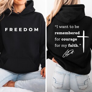 May include: Black hoodie with the word "FREEDOM" in white block letters on the front. The back of the hoodie features a white cross and the quote, "I want to be remembered for courage for my faith."