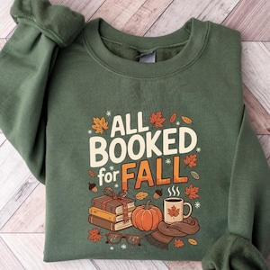 May include: A military green sweatshirt with the text "ALL BOOKED for FALL" in white and orange. The graphic includes books, a pumpkin, a mug, and fall leaves. The sweatshirt is folded on a light wood-grain surface.