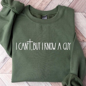 May include: Military green crewneck sweatshirt with the text "I CAN'T, BUT I KNOW A GUY" in white. The sweatshirt is made of a soft material and has ribbed cuffs and a crew neck. The background is a light wood pattern.