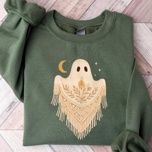 May include: Military green sweatshirt featuring a ghost design with a crescent moon and stars. The ghost has a decorative, fringed shawl. The sweatshirt is made of a soft material and is perfect for Halloween or everyday wear.