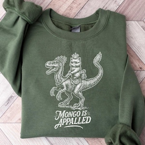 May include: Military green sweatshirt with a white graphic of a cat riding a dinosaur, with the text "MONGO IS APPALLED". The sweatshirt has long sleeves and a crew neck. The background is a light wood pattern.