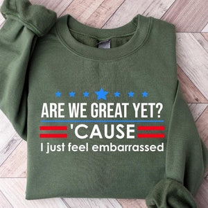 May include: A green sweatshirt with the text "Are we great yet? 'Cause I just feel embarrassed" in white with red and blue accents.