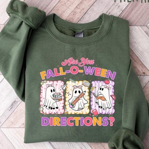May include: Military green sweatshirt with the text "Are You Fall-O-Ween Directions?" and three cartoon ghosts. Each ghost is holding school supplies. The sweatshirt is made of a soft material.