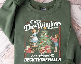 May include: Military green sweatshirt with a crew neck. The front features a vintage illustration of children decorating a Christmas tree, with the text "From The Windows To The Walls, I'm About To Deck These Halls."