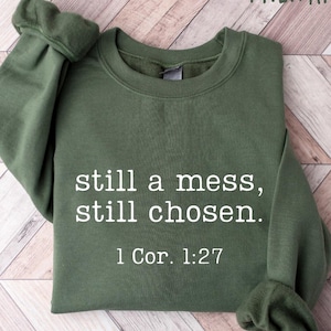 K&ouml;nnte beinhalten: Milit&auml;rgr&uuml;nes Sweatshirt mit dem Text "still a mess, still chosen. 1 Kor. 1:27" in Wei&szlig;. Das Sweatshirt ist auf einer hellen Holzmaserungsoberfl&auml;che gefaltet. Die &Auml;rmel sind umgeschlagen.