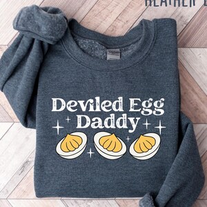 May include: Heather dark gray sweatshirt with the text "Deviled Egg Daddy" in white, along with three deviled egg illustrations. The sweatshirt is folded on a wooden surface.