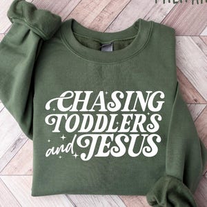 May include: Military green sweatshirt with the text "CHASING TODDLERS and JESUS" in white script. The sweatshirt is made of a soft material and has a crew neck and long sleeves. The sleeves are cuffed at the wrists.