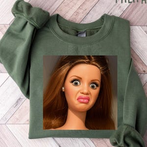 May include: Military green sweatshirt featuring a doll's face with a surprised expression. The doll has brown hair, large eyes, and a puckered mouth. The text "MILITARY GREEN" is printed at the top of the image.
