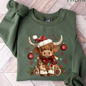 May include: Military green sweatshirt featuring a Highland cow wearing a Santa hat, a red and black plaid scarf, and Christmas lights. The cow is adorned with ornaments and snowflakes, creating a festive holiday design.