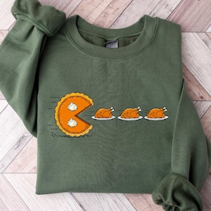 May include: Military green sweatshirt with a graphic of a pie chasing three roasted turkeys. The pie has a bite taken out of it, with whipped cream on top. The text "MILITARY GREEN" is at the top of the image.