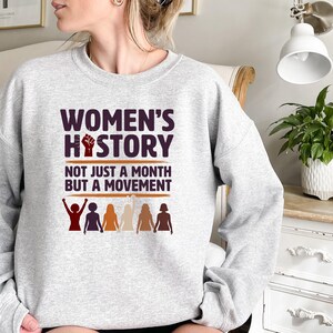 Puede incluir: Una sudadera gris claro con el texto "WOMEN'S HISTORY" en morado oscuro y naranja. Debajo, en blanco, la frase "NOT JUST A MONTH BUT A MOVEMENT". El dise&ntilde;o incluye un pu&ntilde;o en alto y siluetas femeninas.