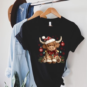 May include: Black t-shirt featuring a festive Highland cow illustration. The cow wears a Santa hat, a plaid scarf, and is adorned with Christmas lights and ornaments. The shirt is hanging on a wooden hanger.