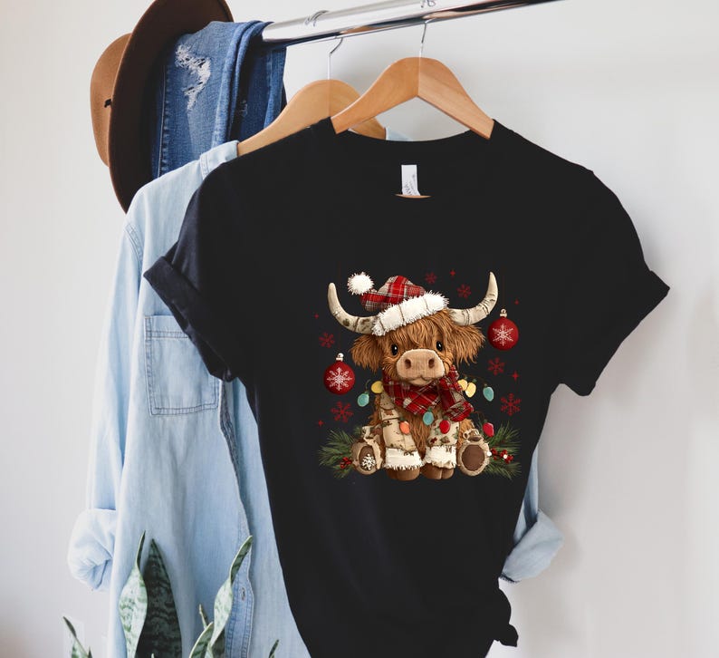 May include: Black t-shirt featuring a festive Highland cow illustration. The cow wears a Santa hat, a plaid scarf, and is adorned with Christmas lights and ornaments. The shirt is hanging on a wooden hanger.