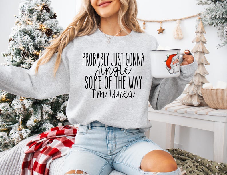 Op de afbeelding: Een lichtgrijs sweatshirt met de tekst "Probably just gonna jingle some of the way I'm tired" in het zwart. Een witte mok met een kerstman-afbeelding wordt in de hand gehouden. Een kerstboom staat op de achtergrond.