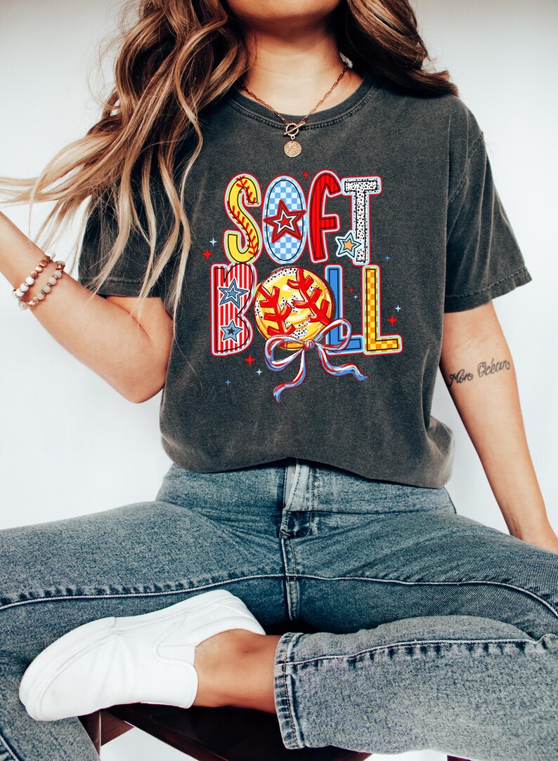 Puede incluir: Una camiseta gris oscuro con un gr&aacute;fico colorido de la palabra "SOFTBALL" con una pelota de b&eacute;isbol y estrellas. La camiseta la lleva puesta una persona con el pelo largo y rubio y unos vaqueros azules.