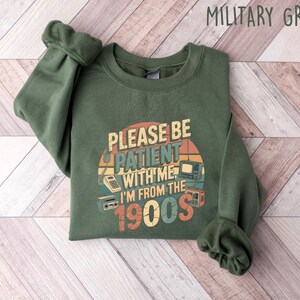May include: Military green sweatshirt with the text "PLEASE BE PATIENT WITH ME I'M FROM THE 1900s" in a retro design. The graphic includes images of a calculator, television, and radio. The sweatshirt is folded on a light wood-grain surface.