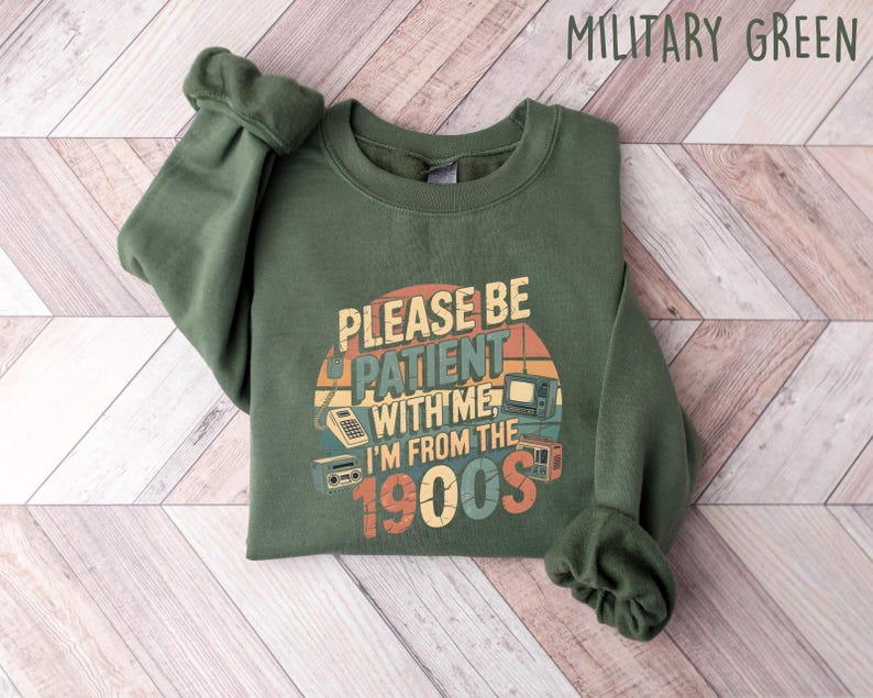May include: Military green sweatshirt with the text "PLEASE BE PATIENT WITH ME I'M FROM THE 1900s" in a retro design. The graphic includes images of a calculator, television, and radio. The sweatshirt is folded on a light wood-grain surface.