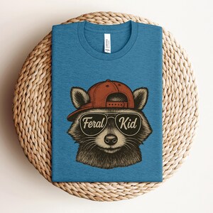 May include: A blue t-shirt featuring a raccoon wearing a red cap and sunglasses. The sunglasses have the words "Feral Kid" printed on them. The shirt is displayed on a woven, circular surface.