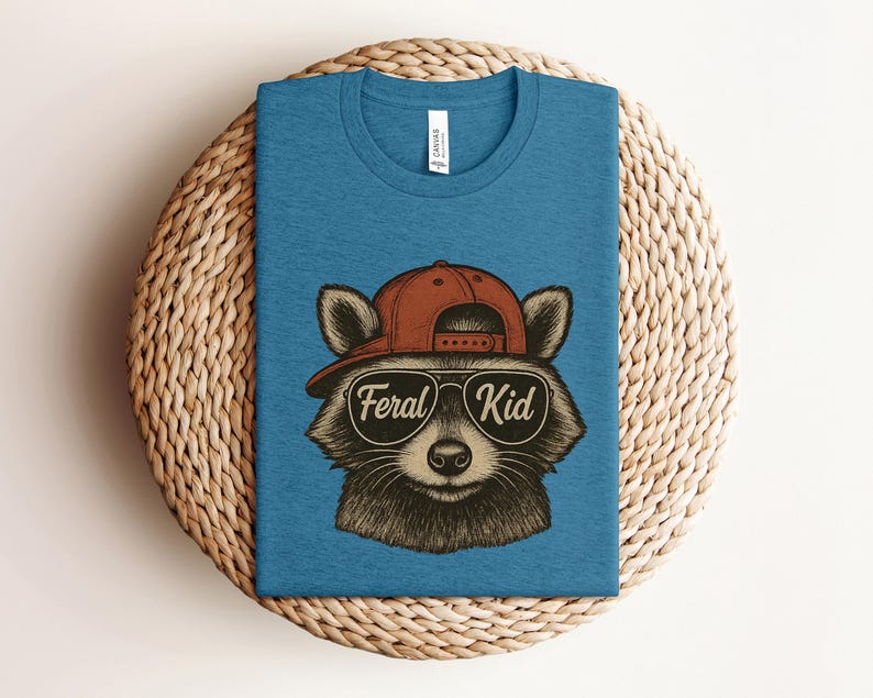 May include: A blue t-shirt featuring a raccoon wearing a red cap and sunglasses. The sunglasses have the words "Feral Kid" printed on them. The shirt is displayed on a woven, circular surface.