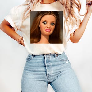 May include: Cream-colored t-shirt featuring a doll's face with a surprised expression. The doll has brown hair, large eyes, and a puckered mouth. The t-shirt is paired with light blue jeans.