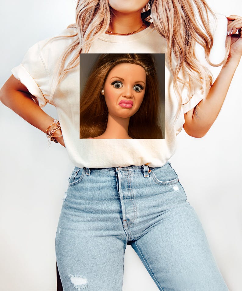 May include: Cream-colored t-shirt featuring a doll's face with a surprised expression. The doll has brown hair, large eyes, and a puckered mouth. The t-shirt is paired with light blue jeans.