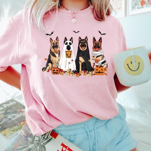 May include: Pink long-sleeve shirt featuring a Halloween-themed graphic. The graphic depicts five German Shepherds dressed in Halloween costumes, including a ghost, mummy, and a dog holding a pumpkin. Black bats and autumn leaves are also included in the design.