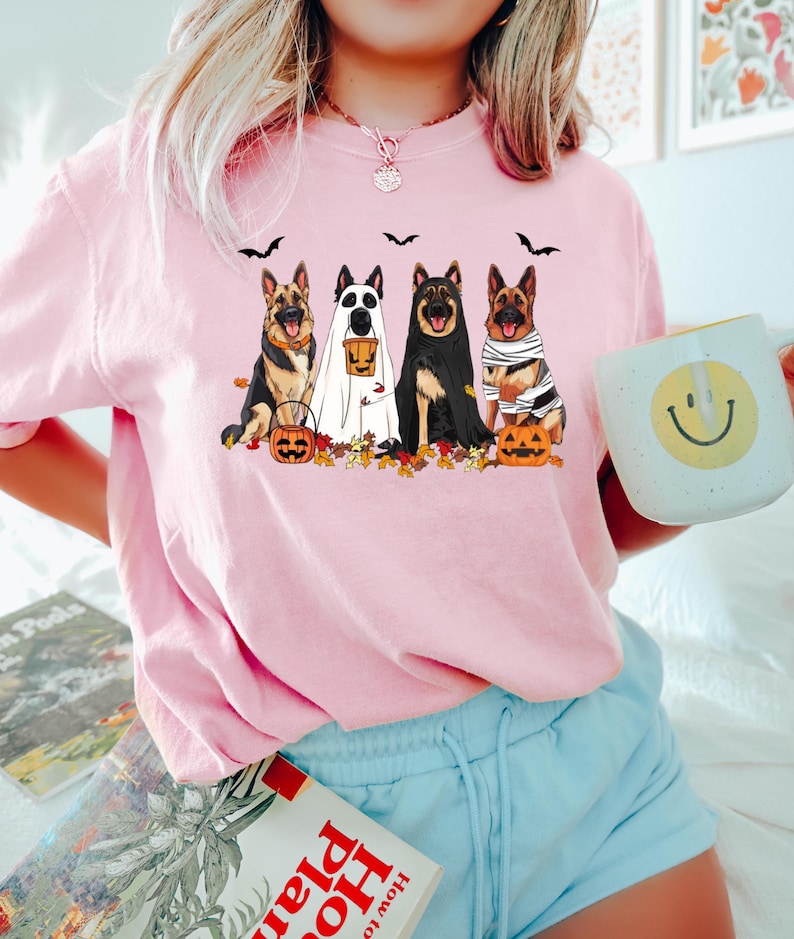 May include: Pink long-sleeve shirt featuring a Halloween-themed graphic. The graphic depicts five German Shepherds dressed in Halloween costumes, including a ghost, mummy, and a dog holding a pumpkin. Black bats and autumn leaves are also included in the design.