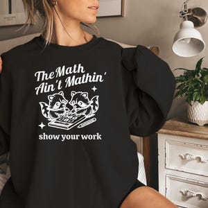 May include: Black sweatshirt with a white graphic of two raccoons using a calculator. The text reads "The Math Ain't Mathin' show your work."