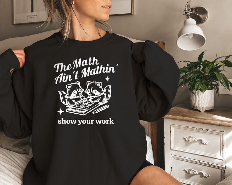 May include: Black sweatshirt with a white graphic of two raccoons using a calculator. The text reads "The Math Ain't Mathin' show your work."