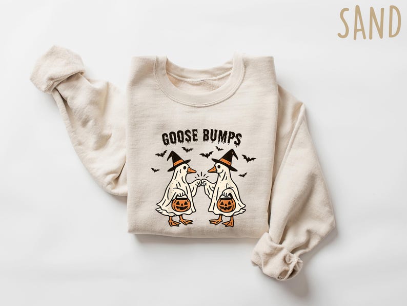 May include: Sand-colored sweatshirt with the text "GOOSE BUMPS" and a Halloween-themed graphic. The graphic features two cartoon geese dressed as ghosts, wearing witch hats, and holding jack-o'-lanterns. Small bats are scattered around the text.