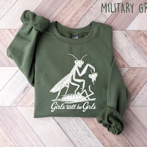 May include: A military green sweatshirt with a white graphic of a praying mantis and the text "Girls will be Girls". The long-sleeved crewneck top is made of a soft material.