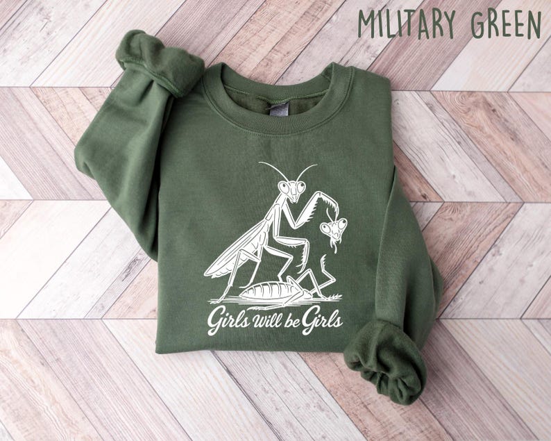 May include: A military green sweatshirt with a white graphic of a praying mantis and the text "Girls will be Girls". The long-sleeved crewneck top is made of a soft material.