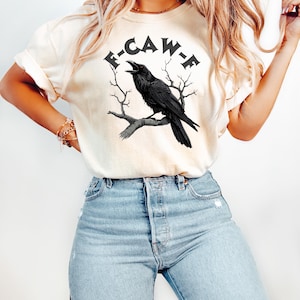 May include: A white t-shirt with a black raven illustration and the text "F-CAW-F" printed on the front.