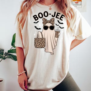 May include: Cream-colored t-shirt with a Halloween-themed graphic. The design features a ghost wearing sunglasses and a bow, holding a leopard print tote bag and a coffee cup. The text "BOO-JEE" is above the ghost, with bat silhouettes.