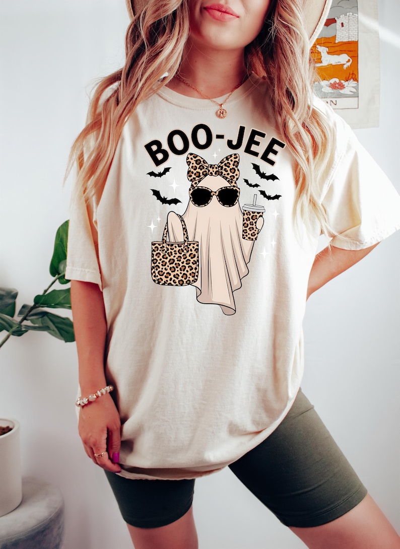 May include: Cream-colored t-shirt with a Halloween-themed graphic. The design features a ghost wearing sunglasses and a bow, holding a leopard print tote bag and a coffee cup. The text "BOO-JEE" is above the ghost, with bat silhouettes.