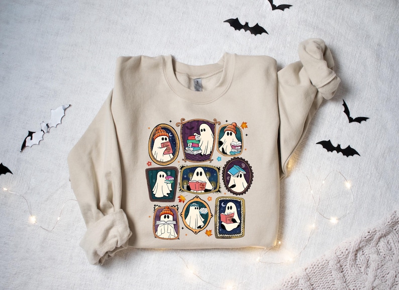 May include: Beige sweatshirt featuring a colorful graphic of cartoon ghosts reading books, each framed in a different ornate style. The ghosts wear hats and scarves, with bats and string lights in the background. A cozy, book-themed design.