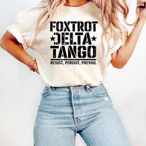 May include: Cream-colored t-shirt with the words "FOXTROT DELTA TANGO" in bold black letters, with stars and the phrase "RESIST, PERSIST, PREVAIL" underneath. The shirt is paired with light blue denim jeans.