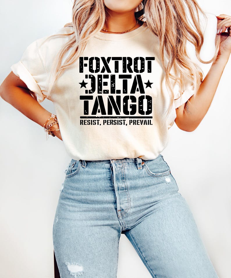 May include: Cream-colored t-shirt with the words "FOXTROT DELTA TANGO" in bold black letters, with stars and the phrase "RESIST, PERSIST, PREVAIL" underneath. The shirt is paired with light blue denim jeans.