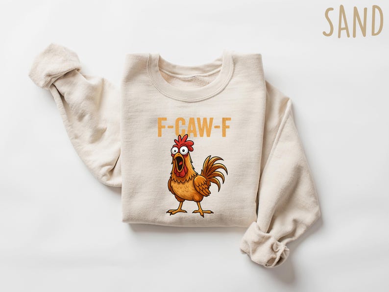 Puede incluir: Una sudadera de color arena con el texto "F-CAW-F" en letras naranjas sobre una ilustraci&oacute;n de un gallo de dibujos animados. El gallo es marr&oacute;n, naranja y rojo con ojos grandes y el pico abierto. Las mangas est&aacute;n enrolladas.