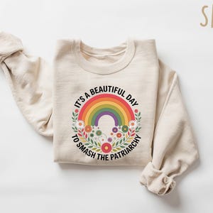 May include: Beige sweatshirt with a colorful rainbow graphic and the text "It's a beautiful day to smash the patriarchy." The rainbow is surrounded by flowers. The word "SAND" is in the upper right corner.