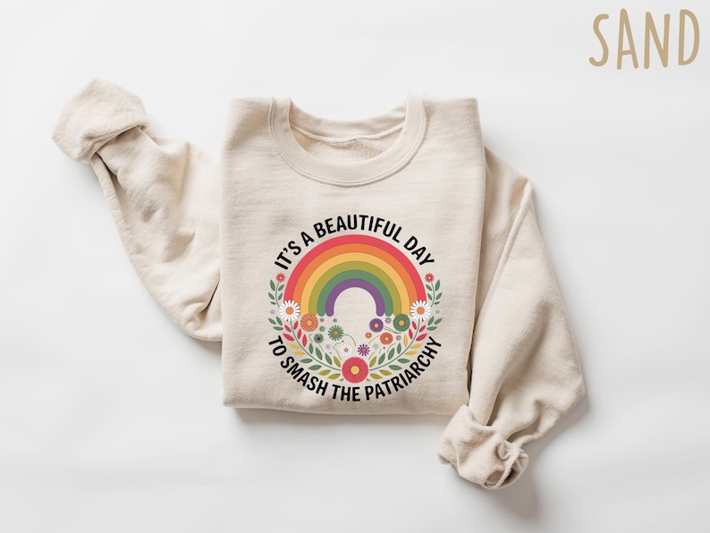 May include: Beige sweatshirt with a colorful rainbow graphic and the text "It's a beautiful day to smash the patriarchy." The rainbow is surrounded by flowers. The word "SAND" is in the upper right corner.