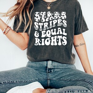 May include: Dark gray t-shirt with the text "STARS STRIPES & EQUAL RIGHTS" in a white, retro font. The word "STARS" has star-shaped patterns. The shirt is paired with blue jeans and white sneakers. A casual, patriotic theme.