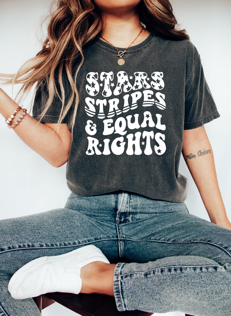 May include: Dark gray t-shirt with the text "STARS STRIPES & EQUAL RIGHTS" in a white, retro font. The word "STARS" has star-shaped patterns. The shirt is paired with blue jeans and white sneakers. A casual, patriotic theme.